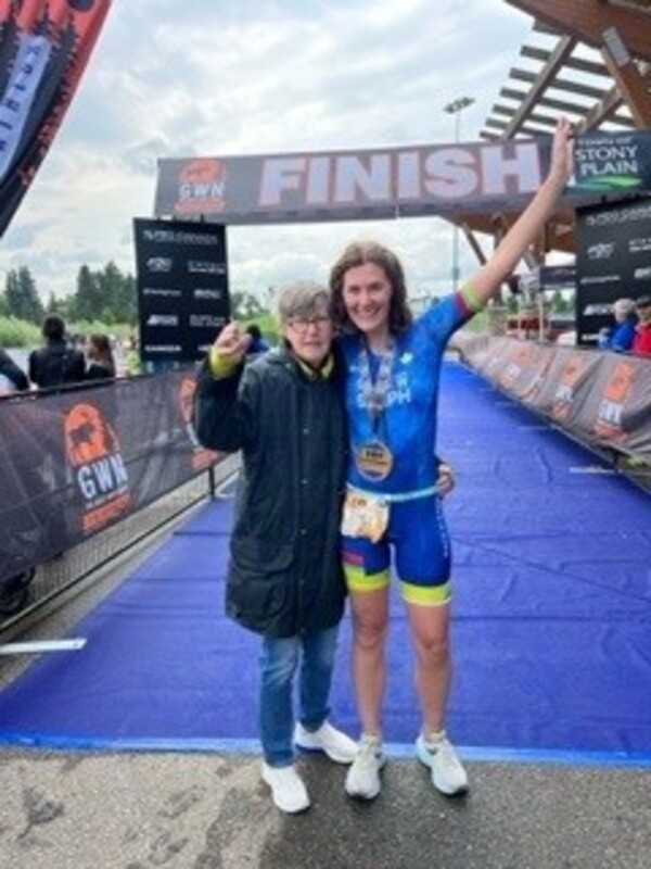 Steph stands at the finish line of her third race, with her arm around her mother. Steph is wearing a blue cycling suit.